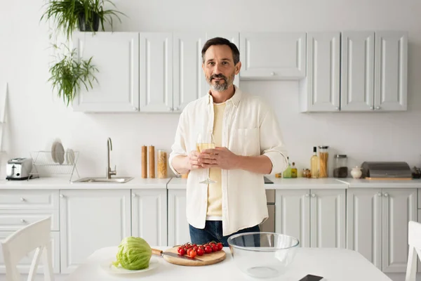 Uomo sorridente con bicchiere di vino bianco guardando la fotocamera vicino a pomodori ciliegini maturi e lattuga fresca — Foto stock