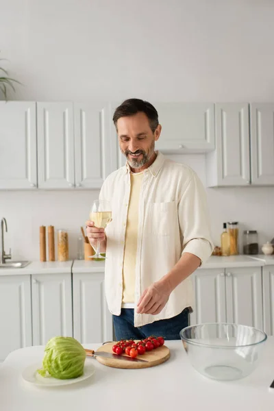 Hombre alegre en camisa blanca sosteniendo copa de vino cerca de lechuga fresca y tomates cherry maduros - foto de stock