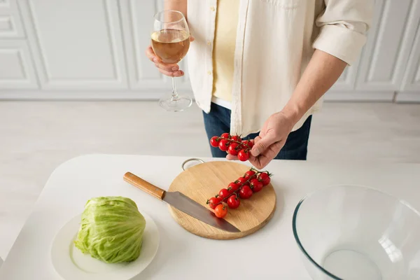 Обрезанный вид человека с бокалом белого вина с помидорами черри возле свежего салата и ножом на разделочной доске — стоковое фото