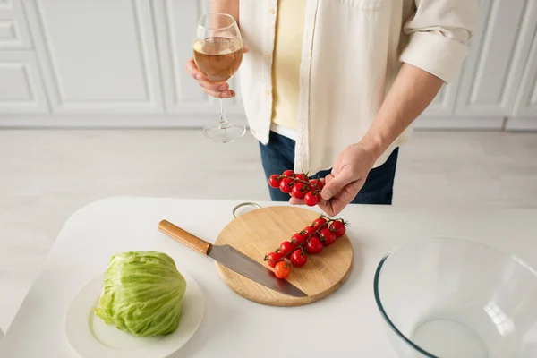 Vista ritagliata dell'uomo con bicchiere di vino bianco che tiene pomodori ciliegini vicino a lattuga fresca e coltello sul tagliere — Foto stock
