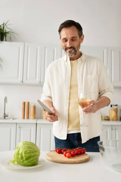Hombre barbudo feliz sosteniendo cuchillo y copa de vino cerca de lechuga fresca y tomates cherry maduros - foto de stock