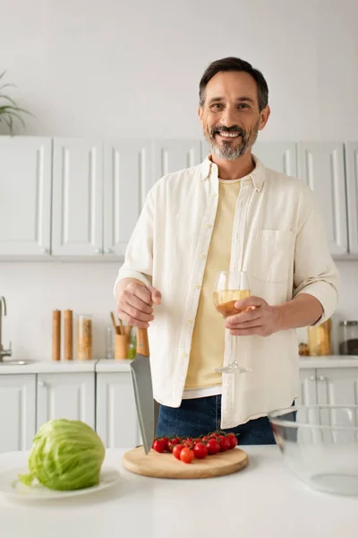 Homme heureux avec couteau et verre de vin regardant caméra près de tomates cerises et de laitue fraîche — Photo de stock