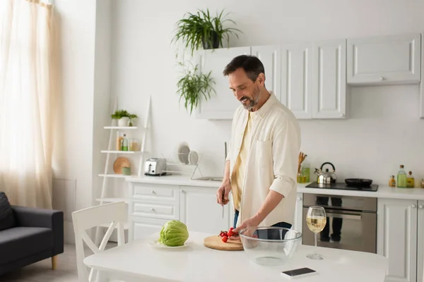 Улыбающийся мужчина держит нож рядом с помидорами черри и свежим салатом на кухне — стоковое фото