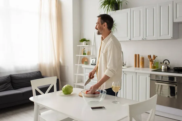 Hombre barbudo feliz mirando hacia otro lado mientras está de pie cerca de verduras frescas y teléfono inteligente con pantalla en blanco en la cocina - foto de stock