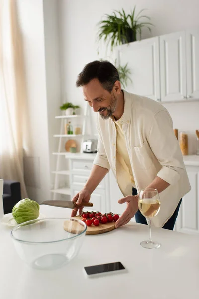 Довольный человек с ножом и нарезанной доской с помидорами черри рядом со свежим салатом и бокалом вина — стоковое фото
