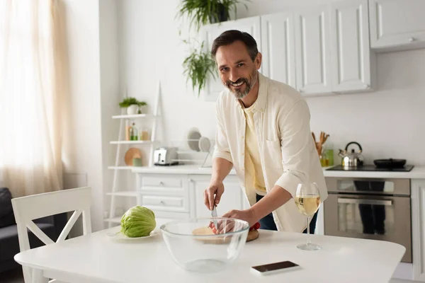 Hombre barbudo sonriendo a la cámara mientras corta tomates cherry cerca de lechuga fresca - foto de stock