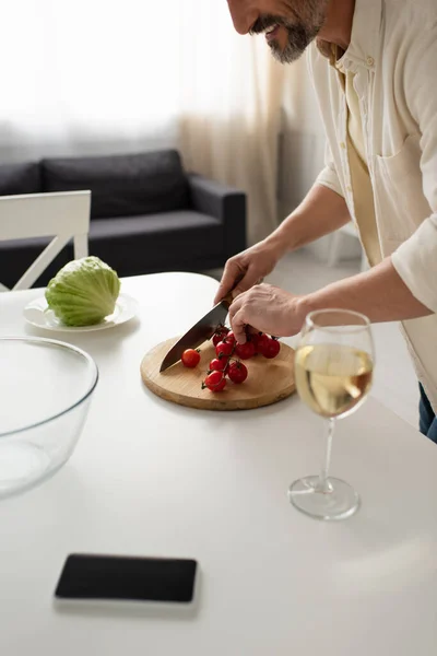 Cropped view of smiling man cutting cherry tomatoes near lettuce and glass of red wine — Stock Photo