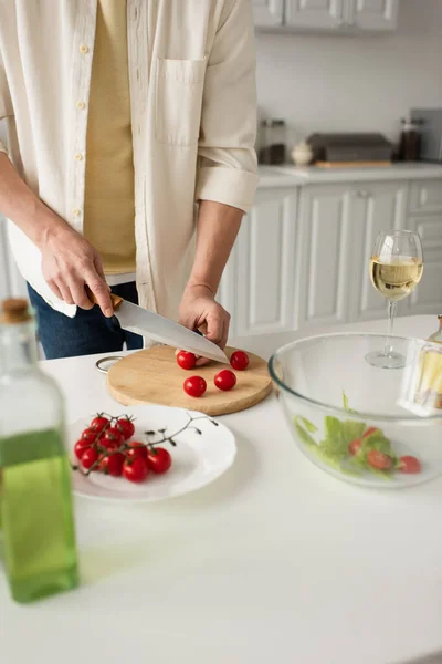 Vista parziale dell'uomo che taglia pomodorini freschi vicino a un bicchiere di vino bianco e una bottiglia di olio sfocata — Foto stock