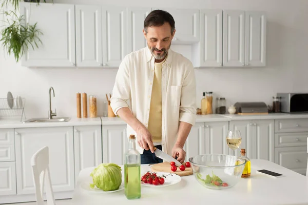 Hombre positivo cortando tomates cherry cerca de lechuga fresca y vaso de vino blanco - foto de stock