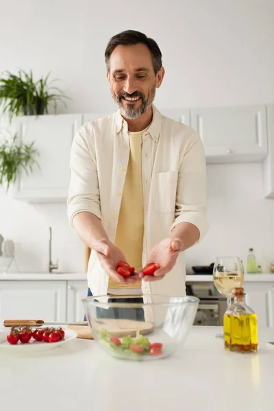 Felice uomo barbuto in possesso di pomodorini tritati vicino ciotola con lattuga e bicchiere di vino bianco — Foto stock