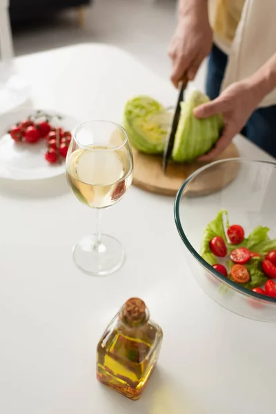 Partial view of blurred man cutting lettuce near bowl with vegetable salad and glass of white wine — Stock Photo