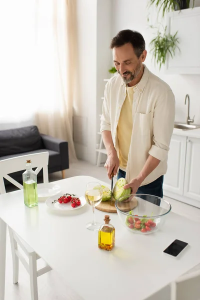 Hombre feliz cortando lechuga cerca de tomates cherry y copa de vino blanco cerca de teléfono inteligente con pantalla en blanco - foto de stock