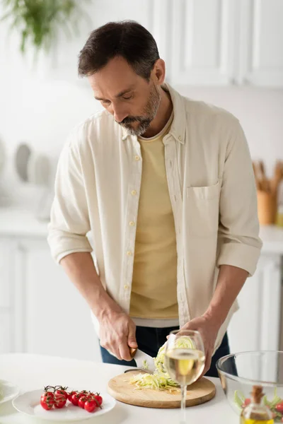 Bearded man in white shirt cutting lettuce near ripe cherry tomatoes and blurred glass of wine — Stock Photo