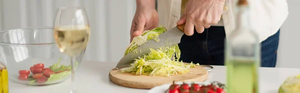 Vista parziale dell'uomo che taglia lattuga fresca vicino alla ciotola con insalata e bicchiere di vino bianco, striscione — Foto stock
