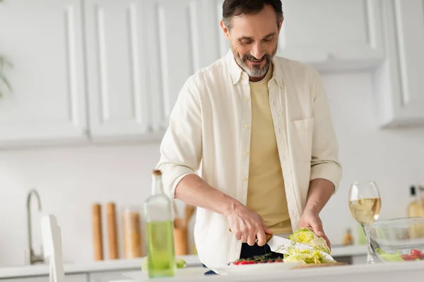 Felice uomo barbuto taglio lattuga vicino bottiglia con olio e bicchiere di vino bianco — Foto stock