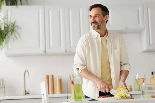 Uomo barbuto sorridente e distogliendo lo sguardo mentre si taglia lattuga vicino al bicchiere di vino bianco — Foto stock