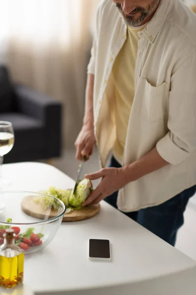 Vista parcial del hombre borroso cortando lechuga fresca cerca del teléfono móvil con pantalla en blanco - foto de stock