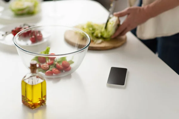 Vue recadrée de l'homme flou coupant la laitue tout en préparant la salade près du téléphone cellulaire avec écran blanc et bouteille d'huile — Photo de stock