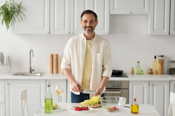 Hombre barbudo sonriendo a la cámara mientras prepara ensalada con lechuga y tomates cherry cerca de un vaso de vino blanco - foto de stock