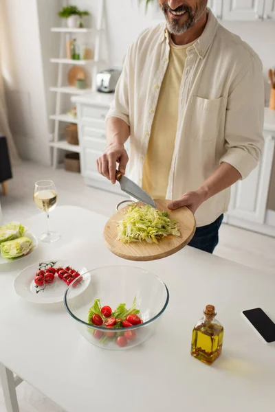 Vista parcial del hombre sonriente preparando ensalada con lechuga y tomates cherry cerca de teléfono inteligente y copa de vino - foto de stock