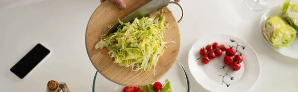 Vue du dessus de l'homme recadré tenant planche à découper avec laitue coupée près de tomates cerises et téléphone portable avec écran blanc, bannière — Photo de stock