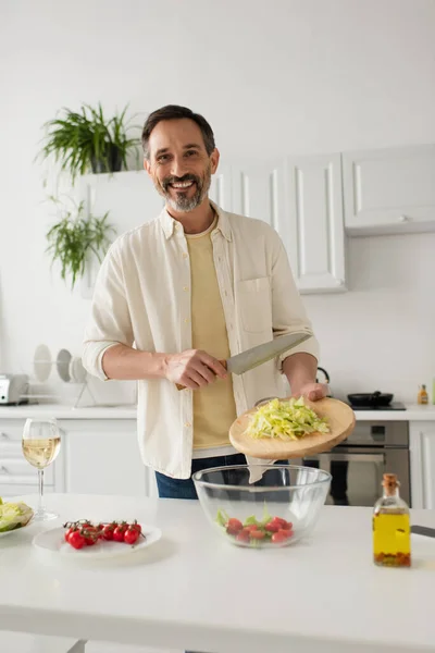 Homem barbudo preparando salada com alface e tomate cereja enquanto sorri para a câmera — Fotografia de Stock