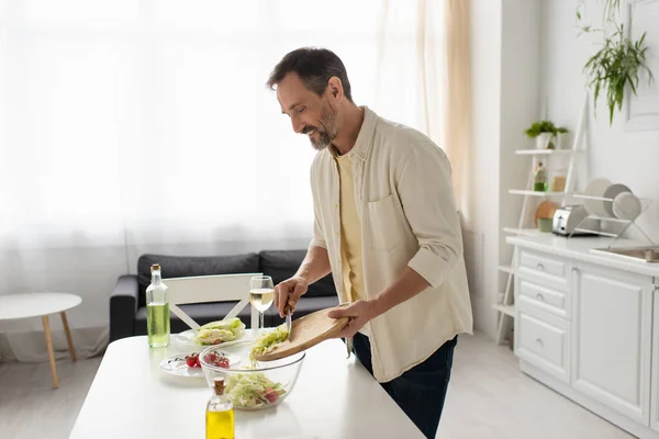 Uomo felice che tiene il tagliere vicino alla ciotola mentre prepara insalata di verdure con lattuga e pomodorini — Foto stock