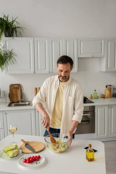 Человек в белой рубашке готовит овощной салат с салатом и помидорами черри возле бокала вина — стоковое фото