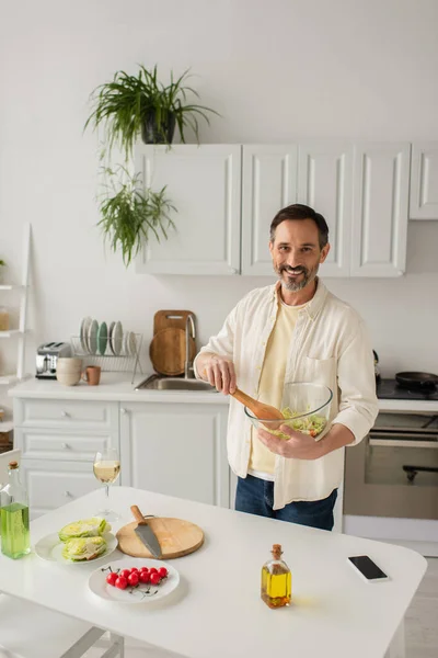 Hombre feliz sosteniendo tazón con ensalada de verduras frescas cerca y copa de vino blanco y teléfono inteligente con pantalla en blanco - foto de stock