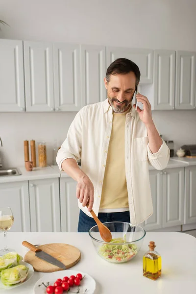 Uomo sorridente in camicia bianca che prepara insalata di verdure fresche e parla su smartphone in cucina — Foto stock