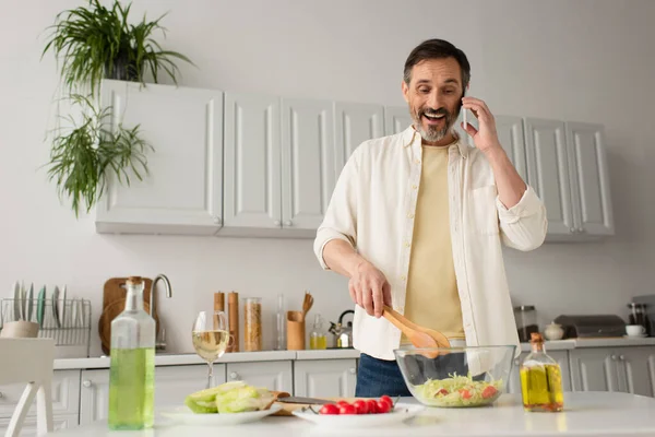 Hombre alegre preparando ensalada de verduras mientras habla por teléfono celular en la cocina - foto de stock