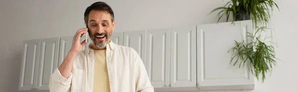 Allegro uomo barbuto che parla al telefono cellulare in cucina, banner — Foto stock