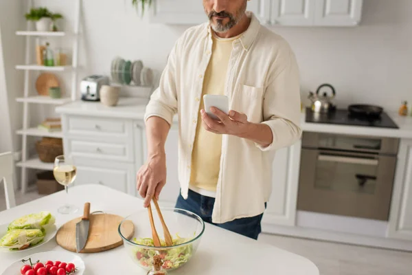 Частичный вид человека, готовящего салат со свежим салатом и помидорами черри, глядя на сотовый телефон — стоковое фото