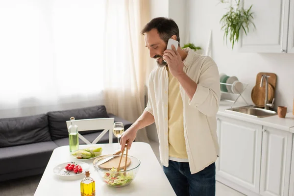 Улыбающийся мужчина разговаривает по смартфону и готовит салат с салатом и помидорами черри — стоковое фото