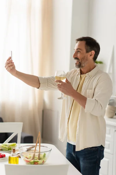 Hombre alegre con copa de vino blanco tomando selfie cerca de verduras frescas y ensalada en la cocina - foto de stock