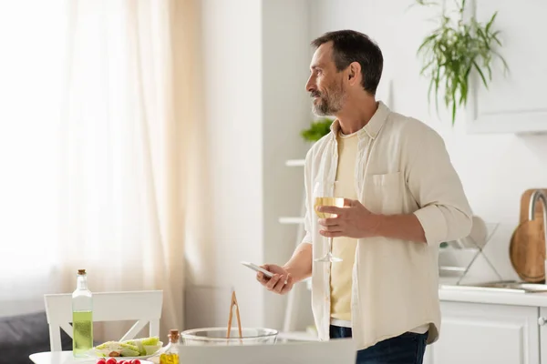 Uomo barbuto con smartphone e bicchiere di vino bianco guardando lontano vicino verdure in cucina — Foto stock