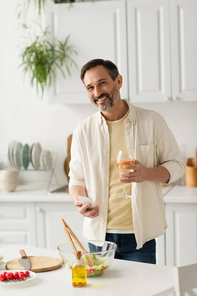 Hombre alegre con teléfono móvil y copa de vino mirando a la cámara cerca de tazón con lechuga y botella de aceite - foto de stock