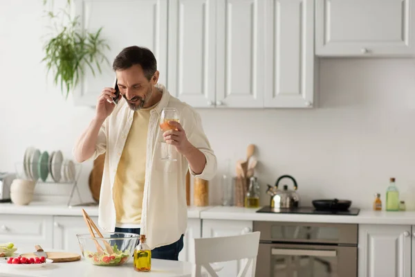 Hombre sonriente con copa de vino hablando en el teléfono inteligente cerca de tomates cherry y tazón con lechuga - foto de stock