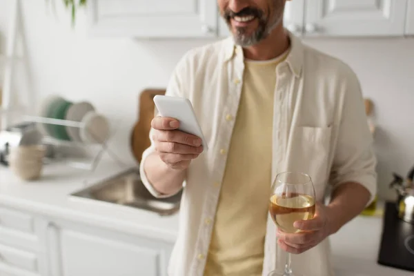 Partial view of smiling bearded man with glass of white wine using smartphone in blurred kitchen — Stock Photo