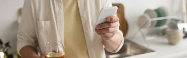 Vista parcial del hombre con copa de vino blanco usando teléfono inteligente en la cocina, pancarta - foto de stock