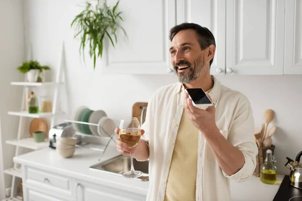 Uomo allegro in possesso di un bicchiere di vino bianco e l'invio di messaggi vocali sul cellulare in cucina — Foto stock