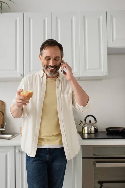 Hombre alegre en camisa blanca hablando por teléfono móvil mientras está de pie con un vaso de vino en la cocina - foto de stock