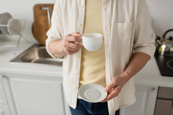 Обрізаний вид людини, що тримає білу чашку кави і блюдце на розмитій кухні — стокове фото