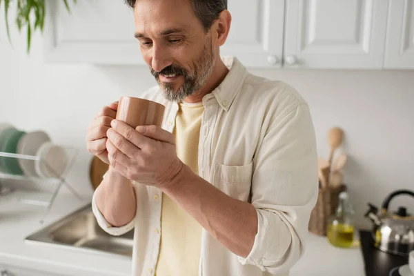 Bärtiger Mann lächelt, während er eine Tasse warmen Tee in der verschwommenen Küche hält — Stockfoto