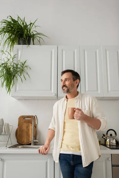 Uomo felice con tazza di tè in piedi vicino lavandino e guardando lontano in cucina — Foto stock