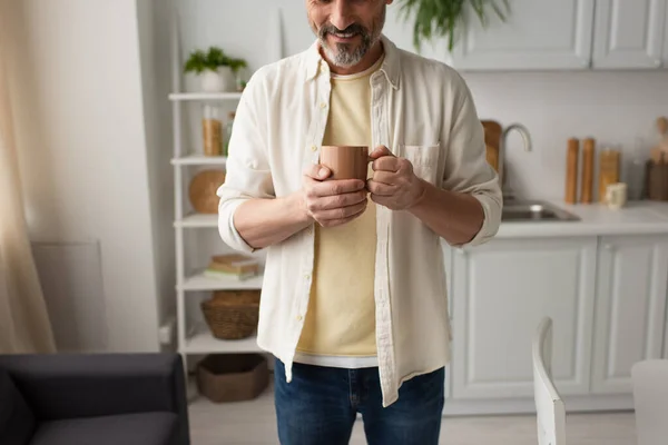 Vista parcial del hombre barbudo sonriente con taza de té caliente en la cocina borrosa - foto de stock