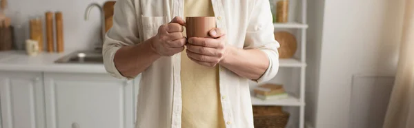 Обрезанный вид человека в белой рубашке держа чашку теплого чая в размытой кухне, баннер — стоковое фото