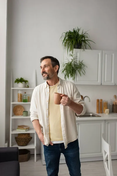 Счастливый бородатый мужчина с чашкой чая стоит с рукой в кармане и смотрит вдаль на кухне — стоковое фото