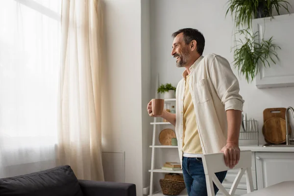 Fröhlicher Mann mit einer Tasse Tee steht am Fenster in der Küche — Stockfoto