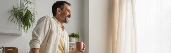 Felice uomo barbuto in camicia bianca in piedi con tazza di tè vicino alla finestra della cucina, banner — Foto stock